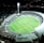 An aerial view of a large, illuminated sports stadium filled with spectators. The stadium is oval-shaped, featuring a green field in the center with markings for a sports game, likely football or rugby. Bright floodlights illuminate the scene, creating a vibrant atmosphere. Surrounding the stadium are urban buildings and streets, barely visible in the night.