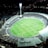 An aerial view of a large, illuminated sports stadium filled with spectators. The stadium is oval-shaped, featuring a green field in the center with markings for a sports game, likely football or rugby. Bright floodlights illuminate the scene, creating a vibrant atmosphere. Surrounding the stadium are urban buildings and streets, barely visible in the night.