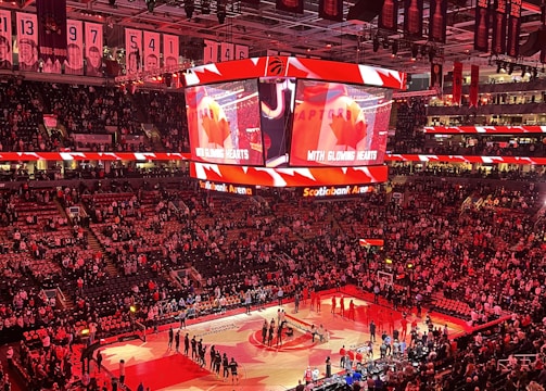 An indoor sports arena filled with spectators and a basketball court illuminated by red lighting. The scoreboards and video screens display promotional content, with numerous banners and championship flags hanging from the ceiling. Players and officials are gathered on the court, possibly in preparation for a game or event.