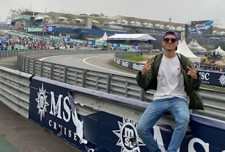 A person in casual attire sits on a barrier beside a racetrack, making peace signs with both hands. In the background, there is a stadium, a large crowd, promotional banners, and a screen displaying an advertisement. The scene has an overcast sky, and the setting appears to be a motorsport event.