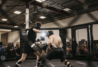 Two fighters engage in a mixed martial arts match within a caged ring. They are wearing protective gear, including gloves and shin guards, and are surrounded by spectators watching intently. Harsh lighting from above casts shadows on the fighters and the mat.