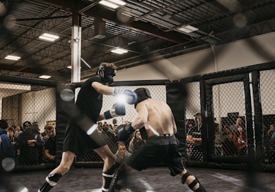 Two fighters engage in a mixed martial arts match within a caged ring. They are wearing protective gear, including gloves and shin guards, and are surrounded by spectators watching intently. Harsh lighting from above casts shadows on the fighters and the mat.