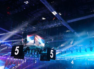A large indoor arena is equipped with advanced lighting and audio systems. Suspended above the space are numerous digital displays showcasing numbers. The environment is filled with blue illumination and spotlights creating a vibrant atmosphere.