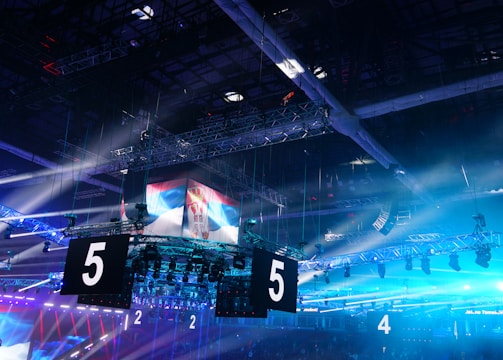 A large indoor arena is equipped with advanced lighting and audio systems. Suspended above the space are numerous digital displays showcasing numbers. The environment is filled with blue illumination and spotlights creating a vibrant atmosphere.
