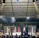 A crowded indoor arena with a cage fight taking place in the center. Audience members surround the cage, with some people involved in organizing the event inside. The arena is well-lit, and the ceiling structure is visible above.